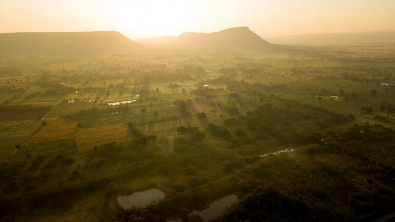 A breathtaking aerial view of a sunlit rural landscape at sunrise, with farmlands and mountains.