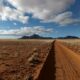 Expansive desert scene with a dirt road leading towards distant mountains under a cloudy sky.