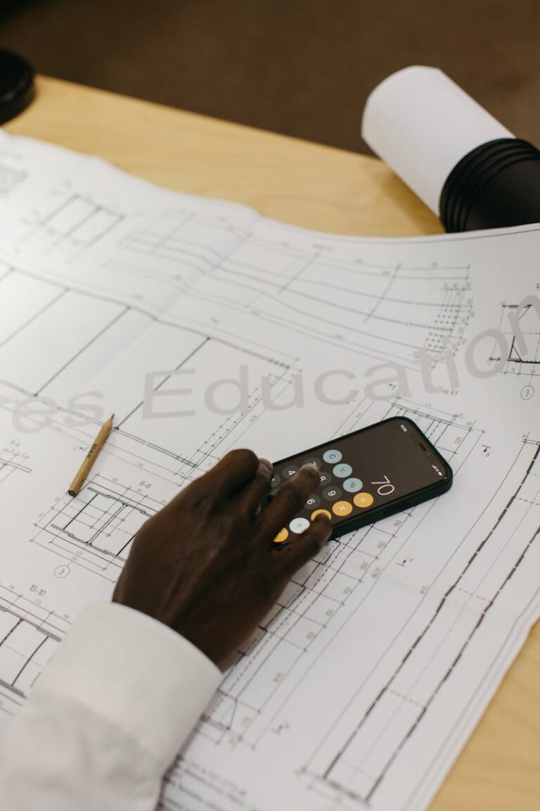 A person using a smartphone for calculations on a detailed architectural blueprint.