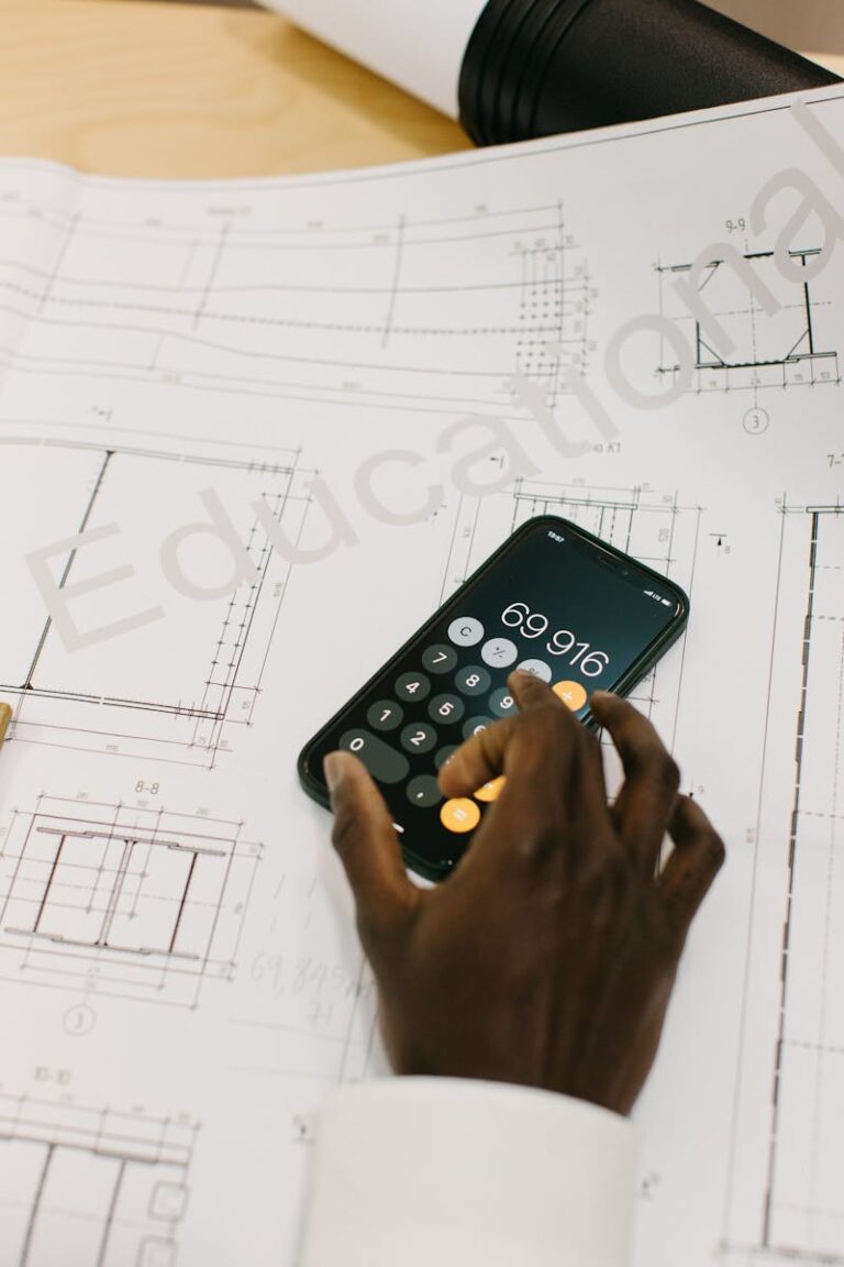 Hand using smartphone calculator on architectural blueprints. Modern technology in engineering.