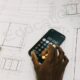 Hand using smartphone calculator on architectural blueprints. Modern technology in engineering.